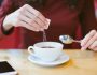 Woman's hands pouring sugar into black coffee - girl sitting at the table with espresso and smartphone - blood and glycemic index control for diabetes -excess of white sugar in food concept