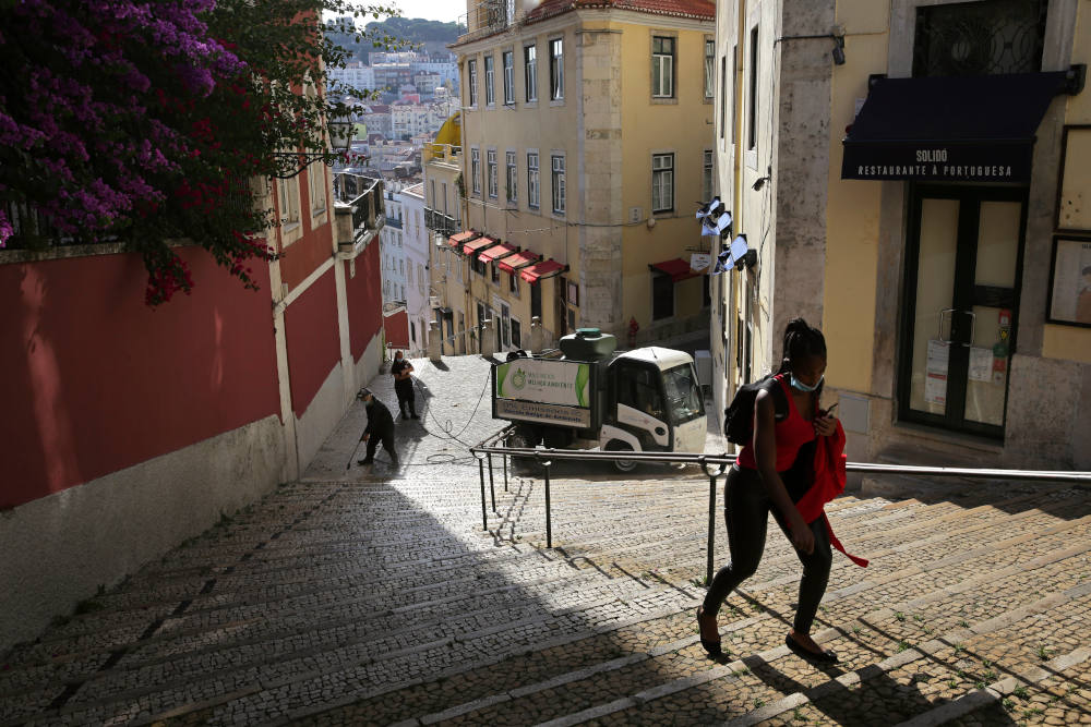 In this photo taken Wednesday, June 24, 2020, a woman wearing a face mask walks past workers washing the street in Lisbon's old center. Portugal avoided the dramatic numbers of infections and deaths recorded by some other European Union countries during the early months of the coronavirus outbreak but since ending its state of emergency and lockdown at the end of April, its total of officially recorded new infections has remained stubbornly high. (AP Photo/Armando Franca)