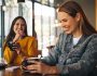 Two women laughing inside a coffee shop. Friends sitting with coffee cups on the table having fun.
