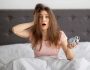 Terrified,Yound,Woman,Holding,Alarm,Clock,In,Bed,At,Home,